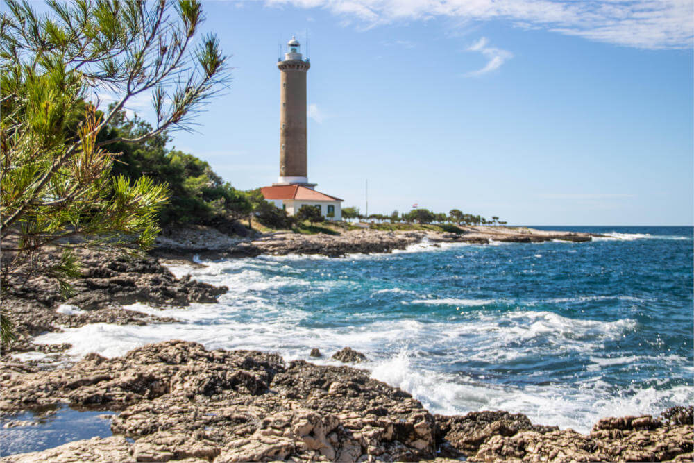 Blick auf den Leuchtturm Veli Rat mit Wellen und Felsen im Hintergrund.